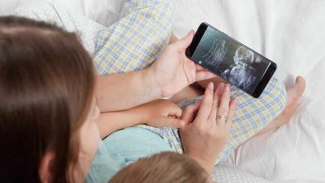 Young mother and little son in pajamas lying in bed and looking on ultrasound image of future baby and brother. Concept of healthcare and family happiness expecting baby