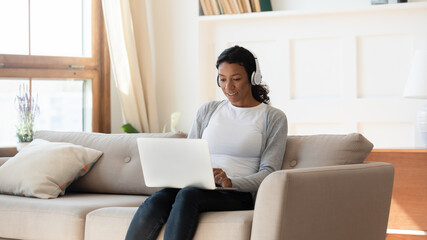 Happy young african american woman in wireless headphones sitting on sofa, looking at laptop screen, involved in distant video call communication with friends or watching educational webinar at home.