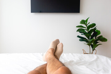 View of man's legs on white bed with TV and plant. Concept of relaxation.