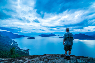 Adventurous male hiker standing on top of a mountain looking at islands in a bay on a cloudy day.