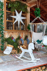Yard of house is decorated for christmas. Xmas decorated terrace of home with big white star on window. Winter porch of house is decorated with fir trees, wooden rocking horse and hanging chair