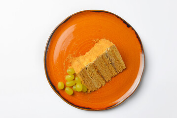 One piece of Honey cake with fresh grapes served on orange plate isolated on white background.