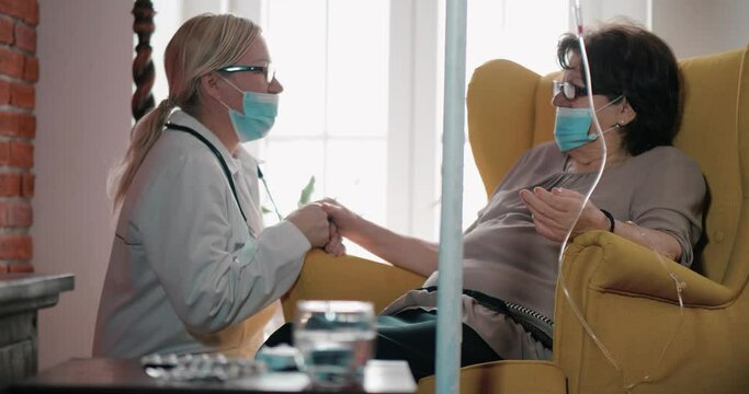 The patient receives an infusion at home. Female nurse talking with elderly woman patient, holding hands. Doctor home visit, checking health of old senior woman during coronavirus, covid-19 quarantine