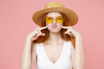 Young redhead woman 20s in straw hat glasses summer clothes point index fingers on blow puff cheeks funny face mouth inflated air crazy expression isolated on pastel pink background studio portrait.