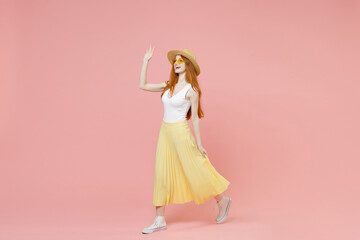 Full length body young smiling redhead ginger woman in straw hat glasses summer clothes maxi skirt walk going waving hand greet someone look aside isolated on pastel pink background studio portrait - obrazy, fototapety, plakaty