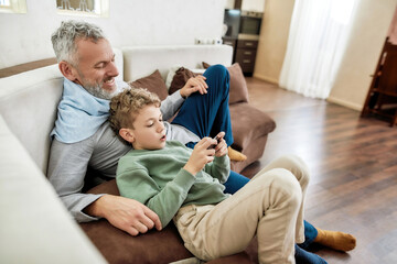 Children and modern technologies. Little boy grandson using smartphone while relaxing on sofa at home with his happy grandfather