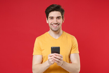 Young smiling unshaved caucasian student man 20s wear casual basic yellow t-shirt holding modern mobile cell phone looking camera reading new surfing isolated on red color background studio portrait