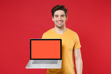 Young smiling unshaved caucasian happy man 20s in yellow t-shirt using holding laptop pc computer with blank screen workspace area copy space mock up isolated on red color background studio portrait.