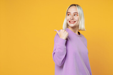 Smiling young blonde woman 20s with bob haircut wearing casual basic purple shirt pointing thumb aside back on workspace area copy space mock up isolated on yellow color background studio portrait