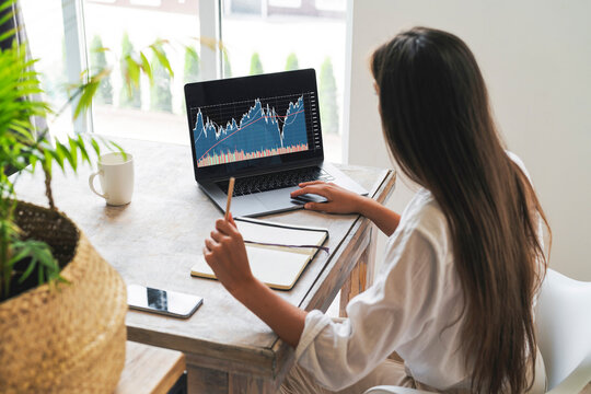 Attractive business woman sits at table in front of laptop and uses smartphone for work. Beautiful brunette girl analyzes stock market while at home. Millennial women on Internet.