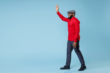 Full length side view joyful young bearded african american man in casual red shirt cap eyeglasses waving and greeting with hand as notices someone isolated on pastel blue background studio portrait.