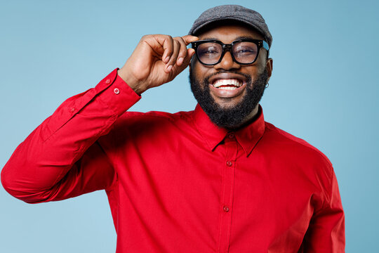 Close up of laughing cheerful young bearded african american man 20s wearing casual red shirt cap eyeglasses standing and looking camera isolated on pastel blue color wall background studio portrait.