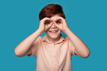 Caucasian ginger boy is gesturing like having binoculars on eyes smiling on a blue studio wall