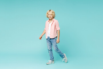 Full length of smiling funny little curly kid boy 10s years old in pink shirt standing looking camera isolated on blue turquoise color background children studio portrait. Childhood lifestyle concept.