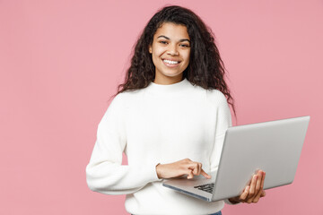 Naklejka premium Young smiling african american woman 20s curly hair wear white casual knitted sweater hold laptop pc computer typing chat on keyboard look camera isolated on pastel pink background studio portrait