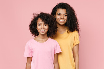 Pretty smiling african american young woman and little kid girl sisters wearing casual t-shirts standing looking camera isolated on pastel pink color background studio portrait. Family day concept.