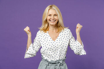 Happy joyful elderly gray-haired blonde woman lady 40s 50s years old wearing white dotted blouse stand doing winner gesture clenching fists isolated on bright violet color background studio portrait.