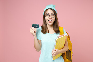Surprised young woman student in casual blue t-shirt hat glasses backpack hold notebooks credit bank card isolated on pastel pink color background. Education in high school university college concept.