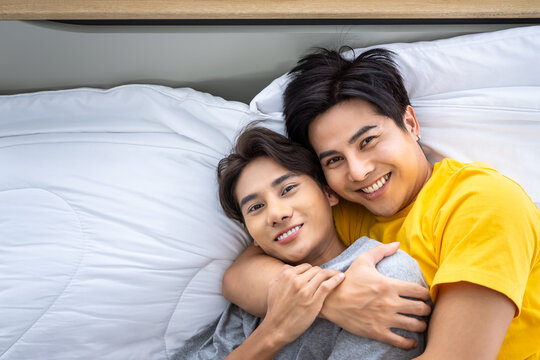 Happy Asian Homosexual Gay Men Male Couple Lying And Embracing On Bed. LGBT And Sexual Diversity Concept. Looking At Camera.