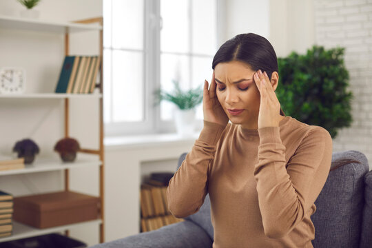 Stressed woman sitting in chair in living-room or office, touching head with grimace of pain, suffering from bad headache, severe migraine, pulsating throbbing pain in temples or temporal arteritis