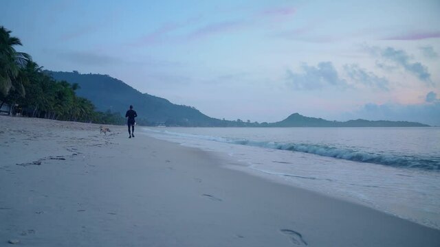 Athletic man in black sportswear running along the beach in the early morning on a tropical island