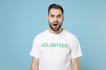 Shocked worried young bearded man in white volunteer t-shirt keeping mouth open looking camera isolated on blue background studio portrait. Voluntary free work assistance help charity grace concept.