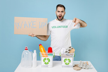 Shocked man in volunteer tshirt stand near recycling stations sorting plastic paper trash hold nameplate wake up showing thumb down isolated on blue background. Voluntary free work assistance concept.