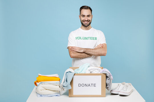 Smiling young man in volunteer t-shirt stand near table with donation box clothes for needy hold hands crossed isolated on blue background. Voluntary free work assistance help charity grace concept.