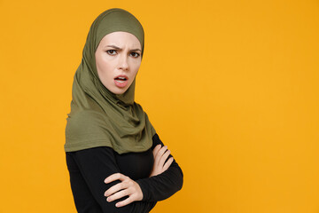 Side view of shocked young arabian muslim woman in hijab black green clothes holding hands crossed looking camera isolated on yellow background, studio portrait. People religious lifestyle concept.