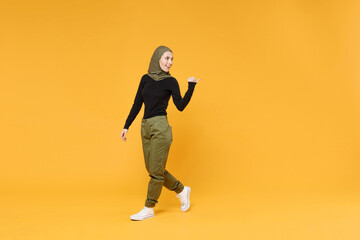 Fototapeta na wymiar Full length side view funny young arabian muslim woman in hijab black green clothes pointing thumb aside isolated on bright yellow color background studio portrait. People religious lifestyle concept.