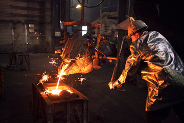 Foundry worker in protective suit and hardhat filling mold with hot molten iron to make parts for...