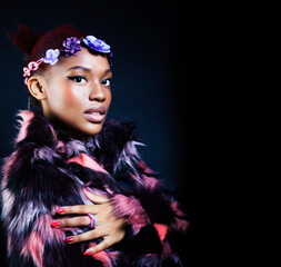 young pretty african american woman in spotted fur coat and flowers on head smiling on black background, fashion people concept