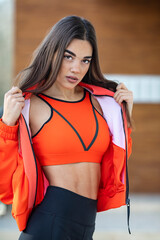 Young sporty woman in modern business district. Relaxed smiling long hair brunette fit woman in tracksuit posing and looking at camera. Young sportswoman