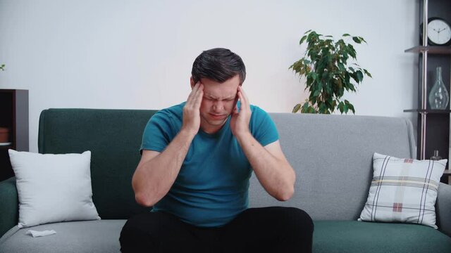 Man suffering from severe headache or migraine, sitting on sofa, young man feeling drunk and pain touching aching head, hangover concept.
