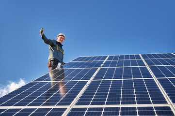 Male worker giving thumbs up and smiling while mounting blue photovoltaic solar panel under beautiful cloudy sky. Concept of alternative energy sources and innovations.