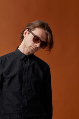 Fototapeta premium Closeup portrait of attractive young man with long hair in sunglasses, wear black shirt, on brown studio background.