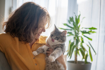 Portrait of a young beautiful woman in a yellow shirt hugging with a gray fluffy cat while sitting...