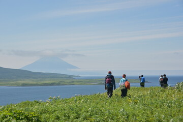 Fototapeta na wymiar Hikers with daypacks walk along green grassy coast on a distant island with big volcano in the background. Expedition to the Kuril islands to the south from Kamchatka peninsula.