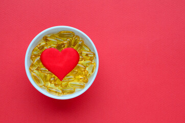 Yellow omega 3 capsules in white bowl and red heart on red background. Concept of proper nutrition, prevention of cardiovascular diseases, Valentine's Day. Medical layout with copy space.