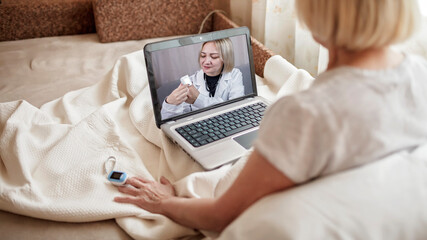 Old woman in bed looking at screen of laptop and consulting with a doctor online at home, telehealth services during lockdown, distant video call, modern tech healthcare application