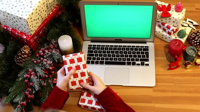 Closeup of young female hands holding present box using laptop keyboard with empty green screen and Christmas decoration on background. Delivery concept. Internet business. Social networking. Mock-up.