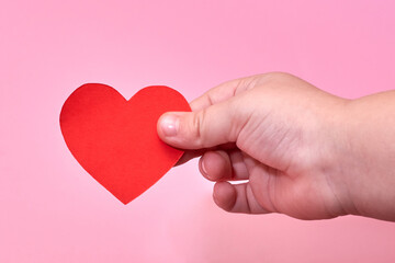 A little boy's hand holds a red paper heart on a pink background.