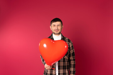 Portrait of a young brunette guy looking to camera, holding a heart-shaped balloon on a red...