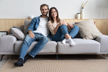 Happy young Caucasian couple relax on cozy sofa at home hugging look in distance dreaming together