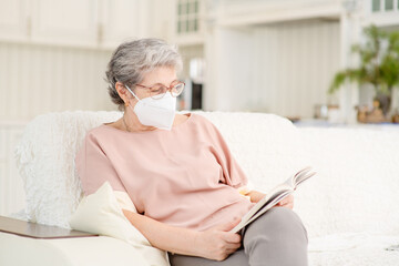 Cute retired old woman with medical mask reading a book while sitting on the couch during the coronavirus lockdown. Ways not to get bored during lockdown and develop while sitting at home.