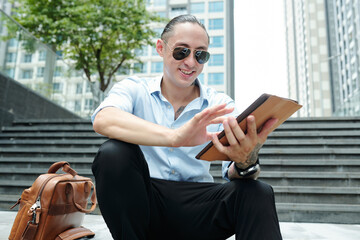 Cheerful man in sunglasses sitting on steps outdoors and using application on digital tablet