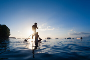 Young happy couple have fun on stand up paddleboard. Active paddle boarder paddling by sunset sea....