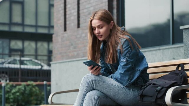 A beautiful young woman is using her smartphone sitting on the bench outside in the city near buildings