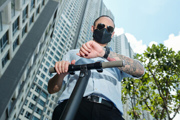 Serious man in sunglasses and protective mask standing on electric scooter and checking notification on smartwatch