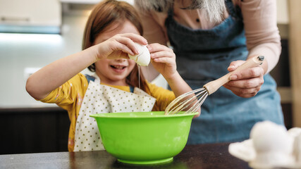 Little girl cracking egg into large bowl near grandmother